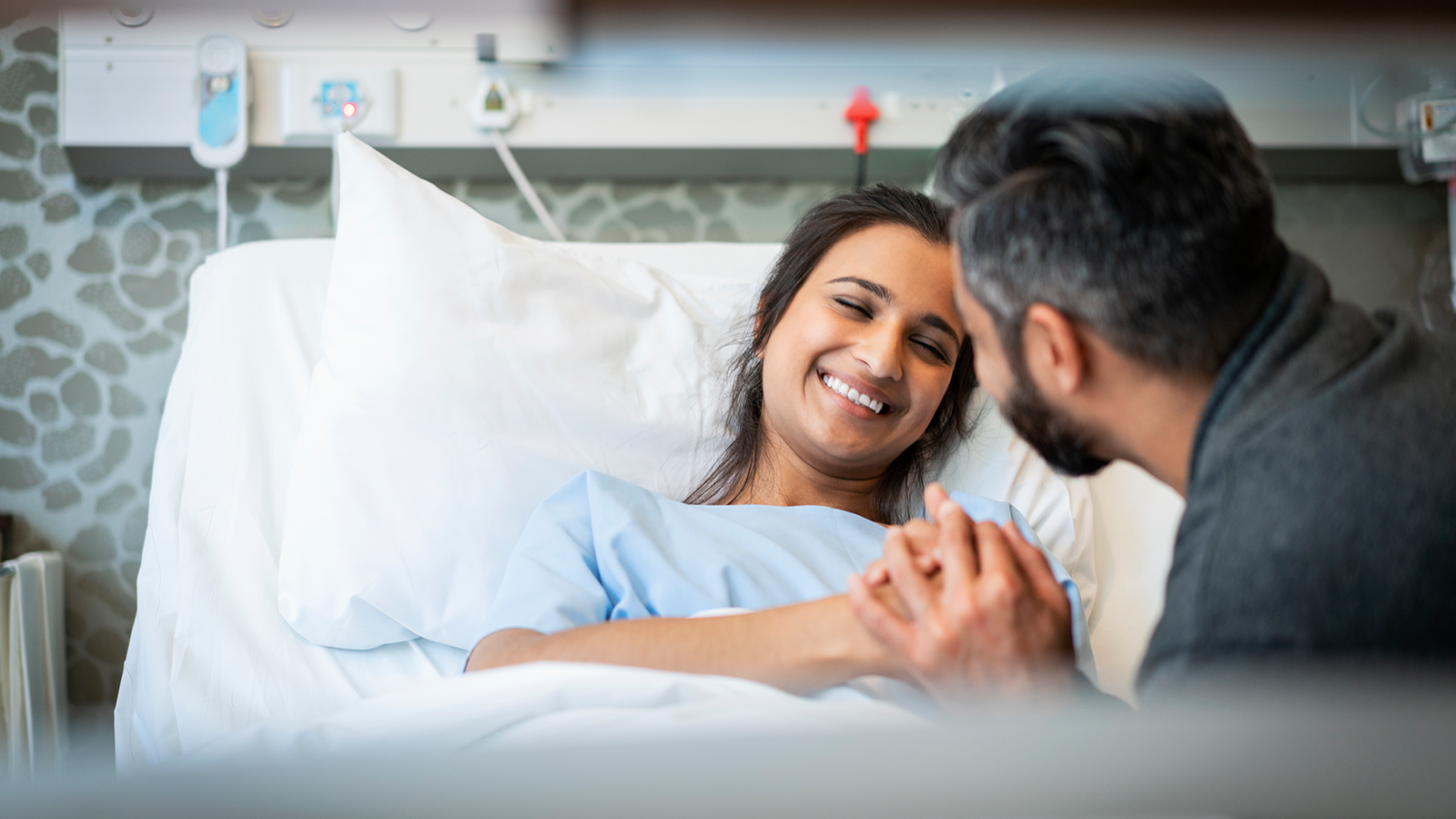 A smiling woman in a hospital gown holds hands with a man leaning towards her, conveying a moment of joy and support in a hospital setting.