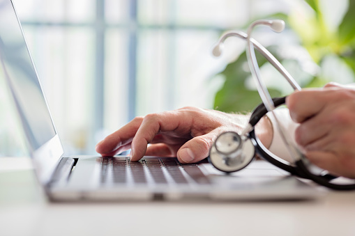 Laptop and two hands with one hand typing and the other holding a stethescope