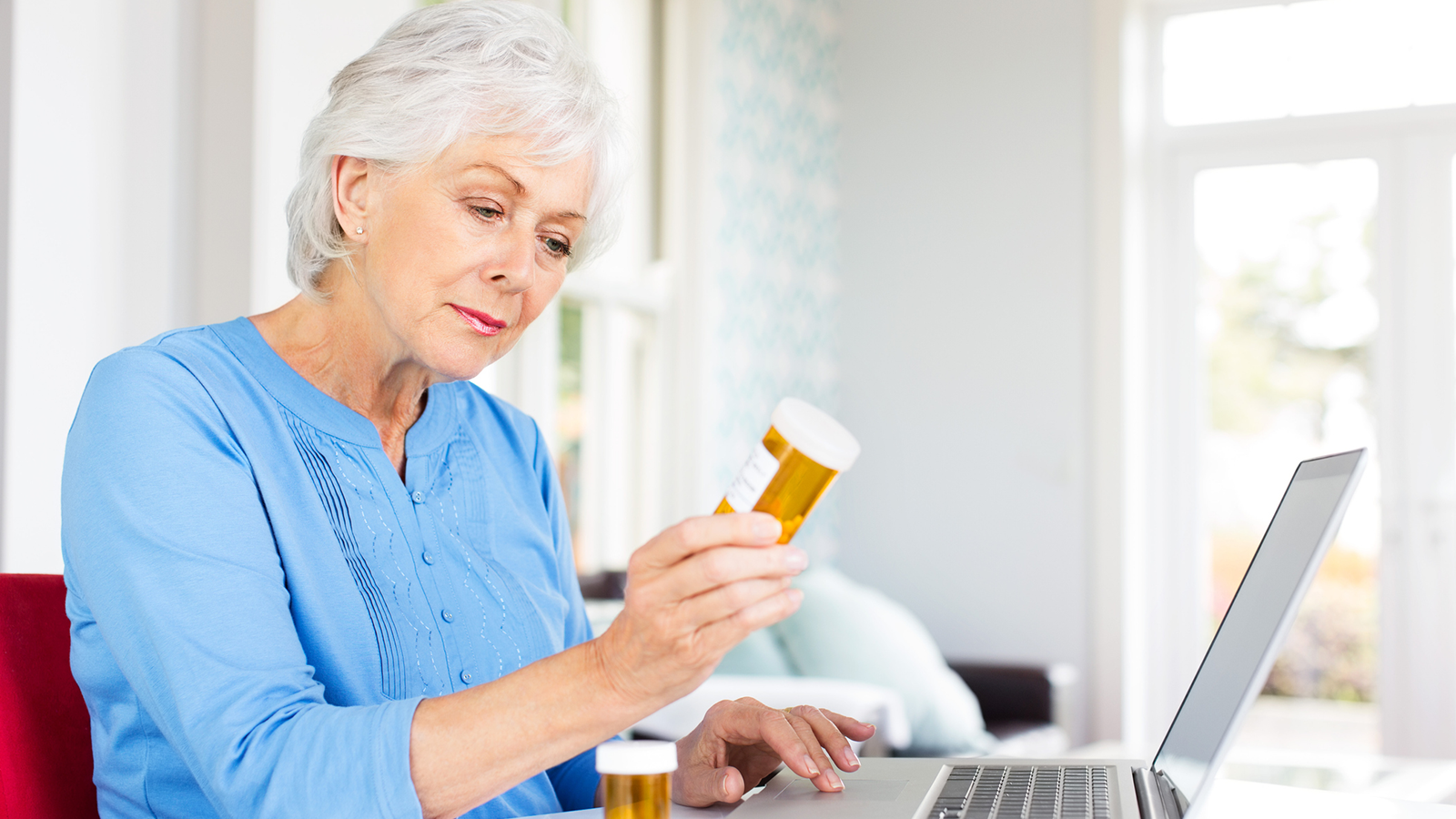 An older woman reviewing a prescription bottle while researching on her laptop at home.